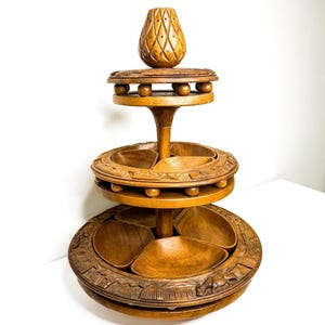 May include: A three-tiered wooden serving stand with a carved design. The stand has three circular trays, each with carved details and divided sections. A decorative wooden piece sits atop the stand. The wood has a warm, natural tone.