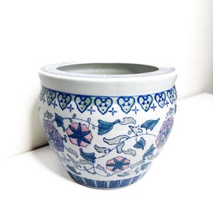 Puede incluir: Una maceta de cerámica blanca con un estampado floral azul, rosa y verde. La maceta tiene forma redonda con una abertura ancha y un borde decorativo. El diseño floral incluye flores y hojas estilizadas.