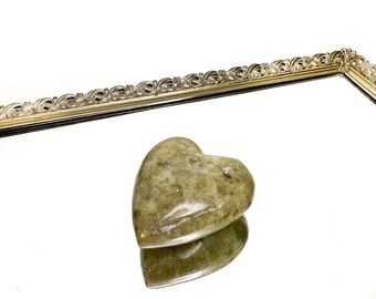May include: A heart-shaped, olive-green stone sits on a mirrored surface, reflecting its form. The stone has a smooth, polished appearance. The mirror is framed by a gold-colored, ornate border, adding a touch of elegance to the composition.
