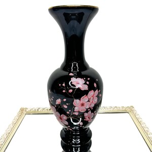 May include: A black vase with a gold rim and a flared top. The vase is decorated with pink cherry blossom flowers and gold accents. The vase is sitting on a mirrored surface.