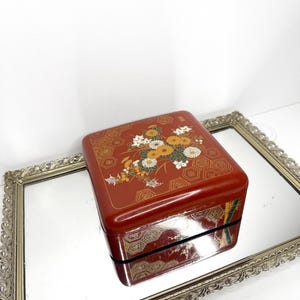 May include: A square, glossy red lacquer box featuring a decorative top panel with gold-toned hexagonal patterns and a central floral arrangement of white and yellow chrysanthemums. The sides of the box also display gold-toned hexagonal motifs.