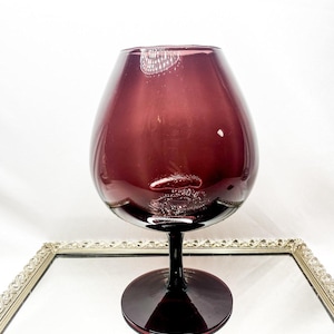 May include: A large, deep purple glass with a rounded bowl and a black stem and base. The glass sits on a mirrored surface within a decorative silver frame. The glass is likely for serving wine or other beverages.