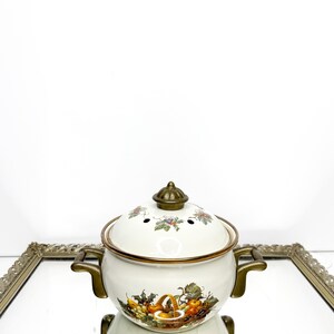 May include: A white enamel pot with a lid, featuring a fruit basket design. The pot has bronze-colored handles and trim. The lid has small holes and a bronze knob. The pot is sitting on a mirrored surface.