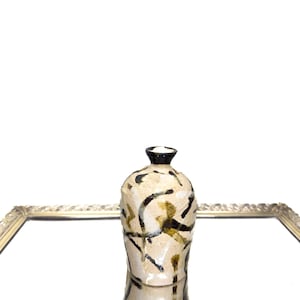 Puede incluir: Un jarrón de cerámica blanca con marcas abstractas negras y marrones. El jarrón tiene un cuerpo redondeado y un cuello estrecho.