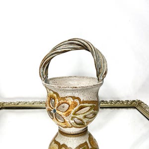 Może przedstawiać: Nakrapiany ceramiczny kosz z plecionym uchwytem. Kosz ma kwiatowy wzór w odcieniach brązu i zieleni. Uchwyt wykonany jest ze skręconych pasm ceramicznych. Kosz odbija się w lustrze.