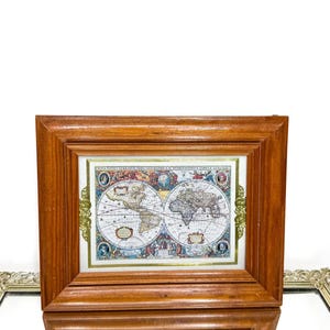 May include: A framed vintage world map print. The map is in a rectangular wooden frame with gold accents. The map depicts the continents and oceans in a detailed, antique style, with decorative elements and text.