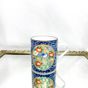 Puede incluir: Una taza de cerámica con asa blanca y diseño azul, dorado y floral. La taza presenta un motivo circular central con un pájaro azul, flores rojas y flores blancas. La taza está colocada sobre una superficie espejada.