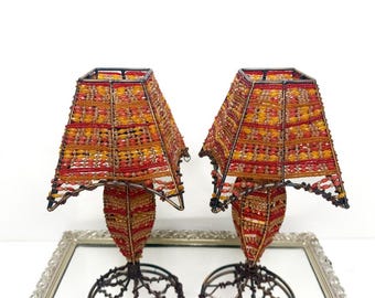 Puede incluir: Dos lámparas decorativas con pantallas hechas de cuentas rojas, naranjas y amarillas. Las lámparas tienen un diseño único con una base de cuentas y una pantalla cuadrada. Las lámparas se exhiben sobre una superficie espejada.