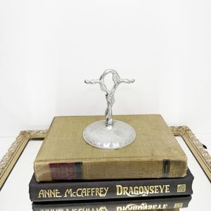 May include: A silver-toned metal stand with a central vertical post that branches into two horizontal arms at the top. The circular base and the post feature embossed grape and vine leaf patterns. The stand is placed on two stacked books.