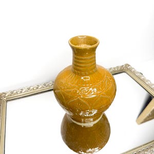 May include: A mustard yellow ceramic vase with a glossy finish, featuring a narrow neck with horizontal ridges and a rounded body decorated with embossed floral and scroll patterns. The vase has a wide opening and a flared rim at the top.