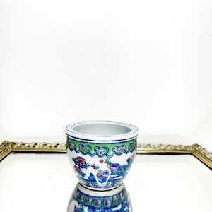 Puede incluir: Una pequeña maceta de cerámica redonda con interior blanco y diseño exterior detallado. La maceta presenta un patrón floral y de pájaros azul, verde y rosa. La maceta se refleja en una superficie espejada.