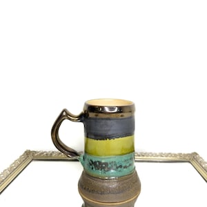 Op de afbeelding: Een handgemaakte keramische mok met een donker handvat en een gouden rand. De mok heeft horizontale banden in zwart, groen en turquoise, met een bruine basis. De mok wordt gepresenteerd op een spiegelend oppervlak.