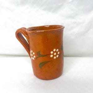 May include: A handmade, terracotta-colored mug with a curved handle. The mug features a floral design with white flower-like shapes and green leaves. The mug is likely made of clay and has a rustic, artisanal appearance.