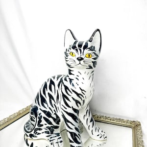 May include: A ceramic cat ornament with a black and white striped design. The cat has yellow eyes and is in a seated position. It is presented on a mirrored tray with a gold-coloured frame. The background is white.