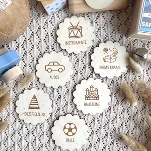May include: Wooden toy set with six scalloped-edge discs, each featuring a different icon and German text. Icons include a drum, car, box of toys, building blocks, a soccer ball, and a stack of blocks. Other wooden toys are in the background.