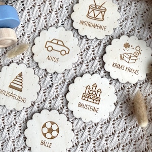 Könnte beinhalten: Runde Holzspielzeug-Etiketten mit gewellten Rändern. Jedes Etikett zeigt eine Spielzeugillustration und Text: "INSTRUMENTE", "AUTOS", "KRIMS KRAMS", "HOLZSPIELZEUG", "BAUSTEINE" und "BÄLLE". für Ikea trofast