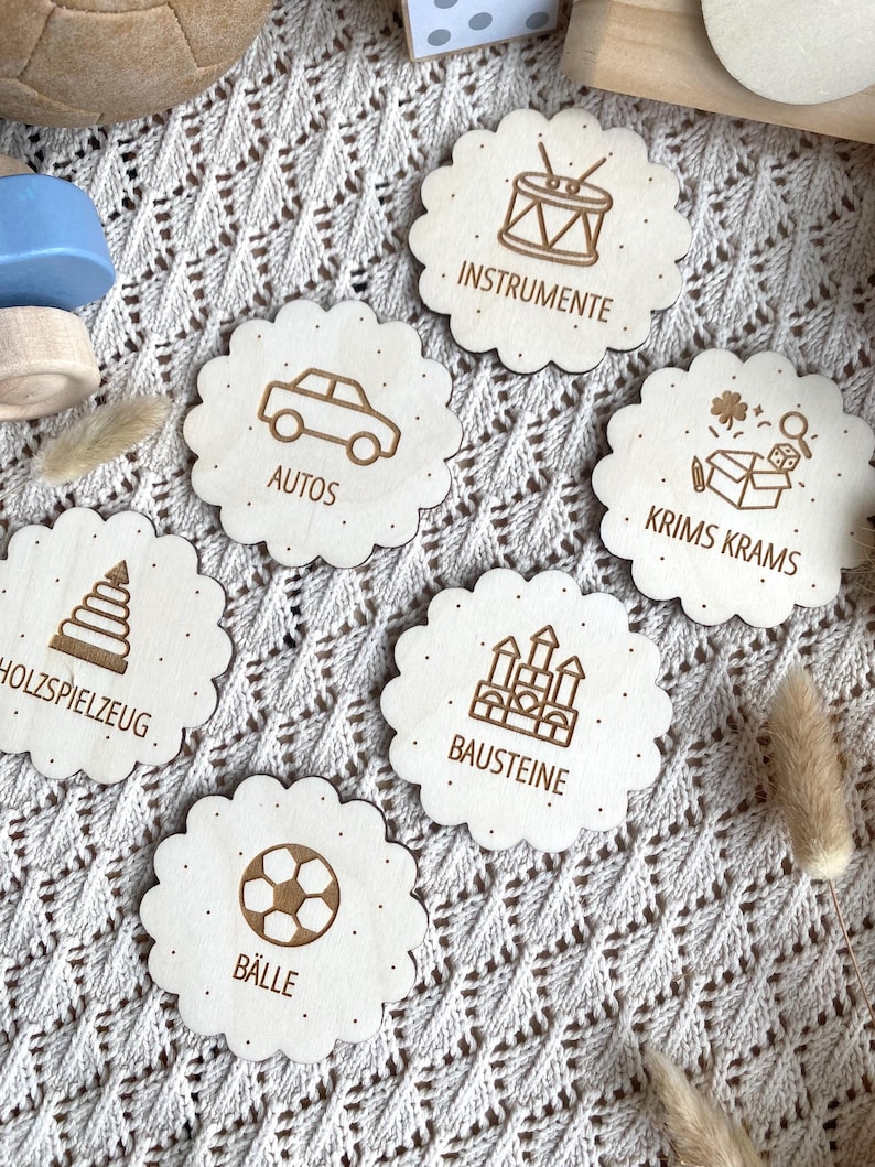 Könnte beinhalten: Runde Holzspielzeug-Etiketten mit gewellten Rändern. Jedes Etikett zeigt eine Spielzeugillustration und Text: "INSTRUMENTE", "AUTOS", "KRIMS KRAMS", "HOLZSPIELZEUG", "BAUSTEINE" und "BÄLLE". für Ikea trofast