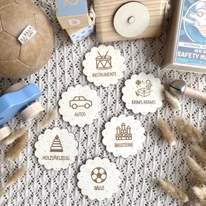 May include: A collection of wooden toys, including a soccer ball, building blocks, and a toy camera. Scalloped wooden discs with engraved illustrations and German words like "Instrumente" and "Autos" are also present. The items are arranged on a textured, off-white surface.