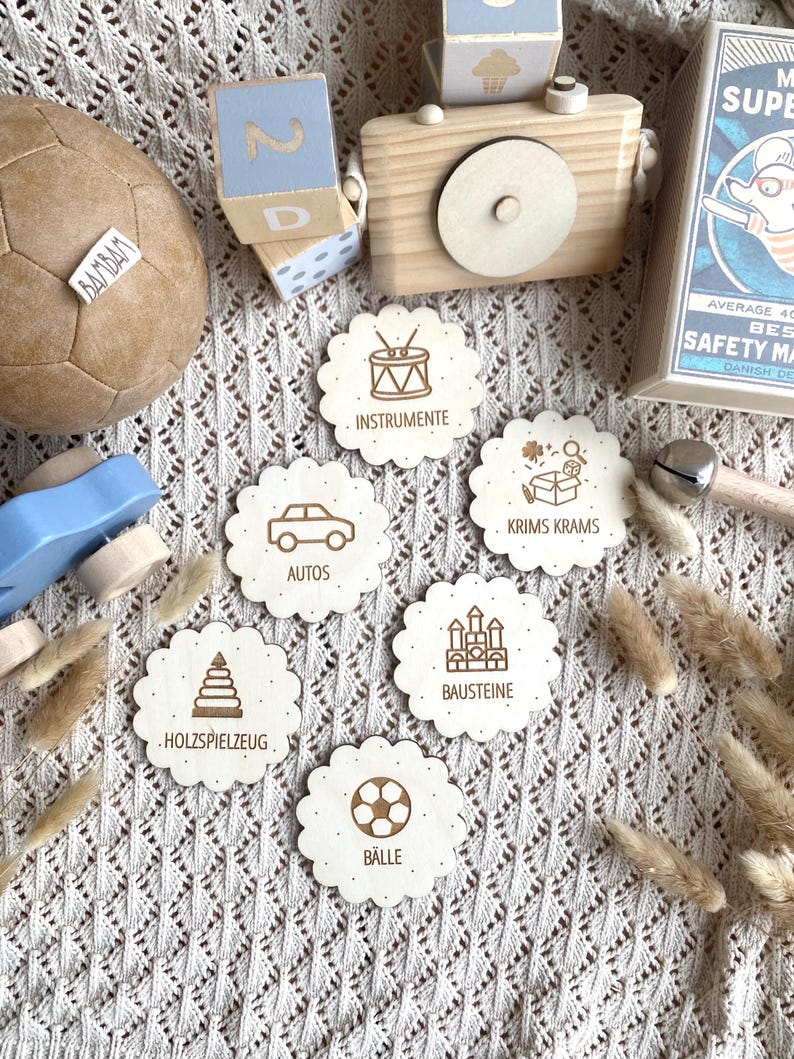 May include: A collection of wooden toys, including a soccer ball, building blocks, and a toy camera. Scalloped wooden discs with engraved illustrations and German words like "Instrumente" and "Autos" are also present. The items are arranged on a textured, off-white surface.