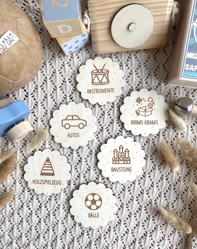 May include: Wooden toy set with six scalloped-edge discs, each featuring a different icon and German text. Icons include a drum, car, box of toys, building blocks, a soccer ball, and a stack of blocks. Other wooden toys are in the background.