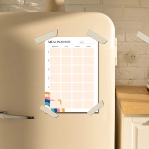 May include: A white meal planner with a colorful abstract design on the left side. The planner is labeled "Meal Planner" and has columns for breakfast, lunch, dinner, and snacks. The planner is attached to a beige refrigerator with tape.