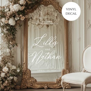 May include: A large, ornate gold mirror with the names "Lilly and Nathan" in elegant script, along with the wedding date. The mirror is surrounded by floral arrangements. A white chair is positioned to the right.
