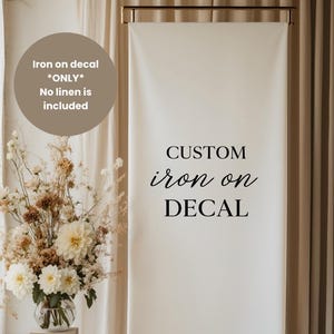 May include: A white banner with the words "CUSTOM iron on DECAL" in black script. A circular graphic above reads "Iron on decal *ONLY* No linen is included". A floral arrangement in a glass vase is in the foreground.