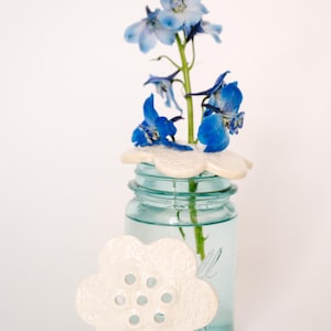 Puede incluir: Un tarro de cristal azul con una tapa de cerámica blanca en forma de flor, que contiene flores azules. Una segunda tapa en forma de flor está colocada junto al tarro. Las flores son de un azul vibrante.