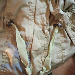 K&ouml;nnte beinhalten: Nahaufnahme einer hellgr&uuml;nen Tarnjacke mit Kordelz&uuml;gen und Metallknebeln. Die Jacke hat ein dezentes Muster aus Gr&uuml;n und Blau, mit sichtbaren N&auml;hten und hellgr&uuml;nen Kordelz&uuml;gen.