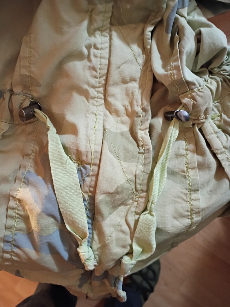 K&ouml;nnte beinhalten: Nahaufnahme einer hellgr&uuml;nen Tarnjacke mit Kordelz&uuml;gen und Metallknebeln. Die Jacke hat ein dezentes Muster aus Gr&uuml;n und Blau, mit sichtbaren N&auml;hten und hellgr&uuml;nen Kordelz&uuml;gen.