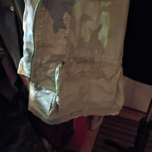 K&ouml;nnte beinhalten: Nahaufnahme einer Jacke mit Tarnmuster, Rei&szlig;verschluss und Kordelzug. Die Jacke hat Gr&uuml;n-, Beige- und Graut&ouml;ne mit hellem Futter. Der Rei&szlig;verschluss hat eine leuchtend gelb-gr&uuml;ne Farbe.