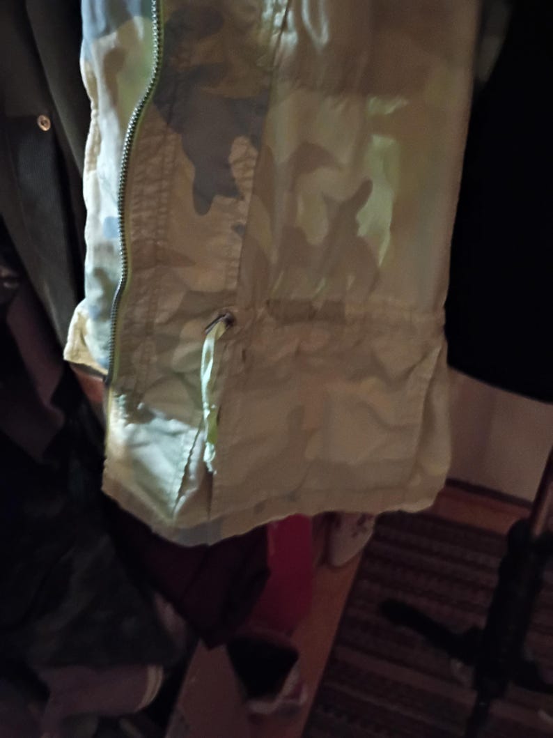 K&ouml;nnte beinhalten: Nahaufnahme einer Jacke mit Tarnmuster, Rei&szlig;verschluss und Kordelzug. Die Jacke hat Gr&uuml;n-, Beige- und Graut&ouml;ne mit hellem Futter. Der Rei&szlig;verschluss hat eine leuchtend gelb-gr&uuml;ne Farbe.