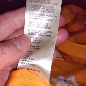May include: A white tag with black text detailing the fabric composition of a garment. The tag reads "Shell 100% Cotton, Lining 100% Rayon, Embroidery 100% Polyester, Coquille 100% Coton, Doublure 100% Rayonne, Broderie 100% Polyester, Deerstoff 100% Baumwolle, Flutter 100% Rayon, Stickerei 100% Polyester, Shell 100% Cotone, Fodera 100% Rayon, Ricamo 100% Poliestere, Exterior 100% Algodon, Forro 100% Rayon, Bordado 100% Poliester, 100% 棉, 100% 人造丝, 100% 锦, In India, Ho en India, 46170, 32054, OR EU, URBN UK Ltd, 146 Brick Lane, LONDON E1 6RU, OP683545".