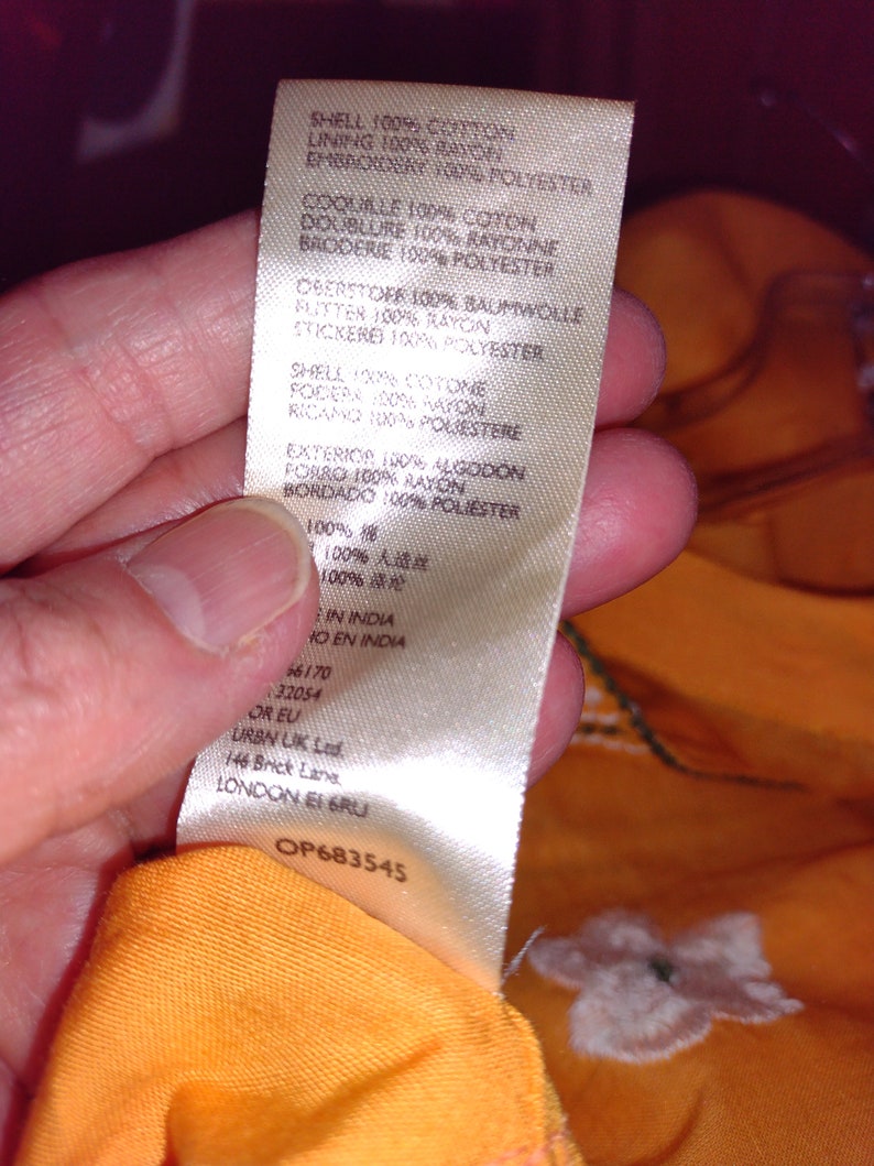 May include: A white tag with black text detailing the fabric composition of a garment. The tag reads "Shell 100% Cotton, Lining 100% Rayon, Embroidery 100% Polyester, Coquille 100% Coton, Doublure 100% Rayonne, Broderie 100% Polyester, Deerstoff 100% Baumwolle, Flutter 100% Rayon, Stickerei 100% Polyester, Shell 100% Cotone, Fodera 100% Rayon, Ricamo 100% Poliestere, Exterior 100% Algodon, Forro 100% Rayon, Bordado 100% Poliester, 100% 棉, 100% 人造丝, 100% 锦, In India, Ho en India, 46170, 32054, OR EU, URBN UK Ltd, 146 Brick Lane, LONDON E1 6RU, OP683545".