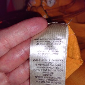 May include: A white care label with instructions for washing and ironing a garment. The label says "Hand wash cold separately, do not wring or twist, use only non-chlorine bleach, line dry, low iron as needed, may be dry cleaned". The label also has French and Japanese text.
