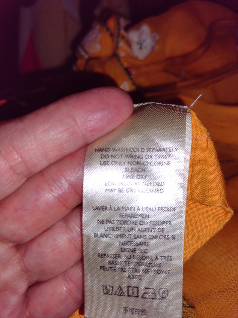 May include: A white care label with instructions for washing and ironing a garment. The label says "Hand wash cold separately, do not wring or twist, use only non-chlorine bleach, line dry, low iron as needed, may be dry cleaned". The label also has French and Japanese text.