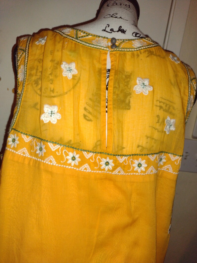 May include: A yellow sleeveless dress with white embroidered flowers and a green trim. The dress has a back zipper and is made of a sheer fabric.