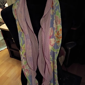 May include: A floral scarf with a light purple gingham pattern on one side and a floral print on the other. The scarf is draped over a black mannequin, showcasing its design and texture. The floral print includes pink, orange, and green flowers.