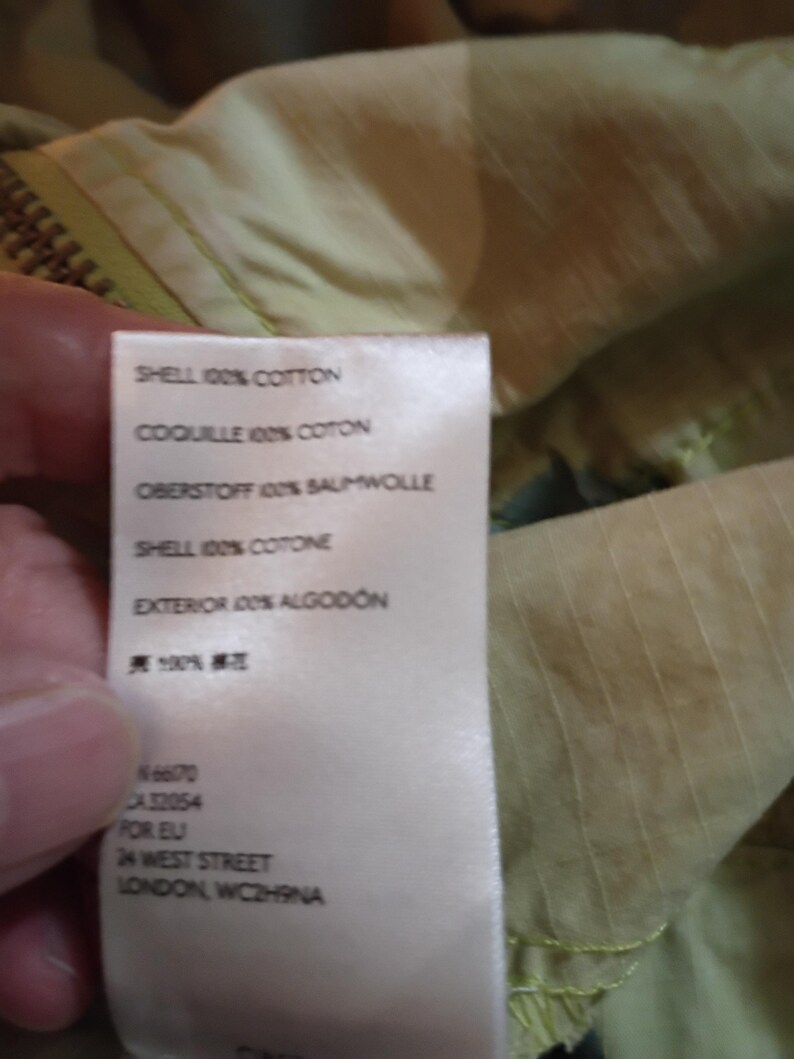 K&ouml;nnte beinhalten: Nahaufnahme eines Stoffetiketts mit Text in mehreren Sprachen, darunter "SHELL 100% COTTON" und "EXTERIOR 100% ALGODON." Das Etikett enth&auml;lt auch die Adresse "24 WEST STREET LONDON, WC2H9NA."