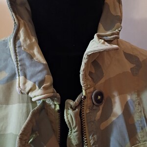 K&ouml;nnte beinhalten: Nahaufnahme einer Tarnjacke mit hohem Kragen und Rei&szlig;verschluss. Die Jacke hat ein Muster aus gr&uuml;nen, beigen und grauen Formen. Ein einzelner Knopf ist in der N&auml;he des Kragens sichtbar.