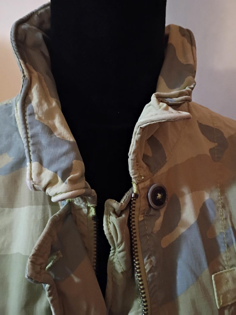 K&ouml;nnte beinhalten: Nahaufnahme einer Tarnjacke mit hohem Kragen und Rei&szlig;verschluss. Die Jacke hat ein Muster aus gr&uuml;nen, beigen und grauen Formen. Ein einzelner Knopf ist in der N&auml;he des Kragens sichtbar.
