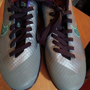 May include: Gray and black athletic shoes with dark purple laces. The shoes have a textured pattern and a teal Nike swoosh logo on the side. The interior is pink and purple.