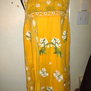 May include: A sleeveless yellow dress with white floral embroidery and green trim. The dress has a round neckline and a loose fit.