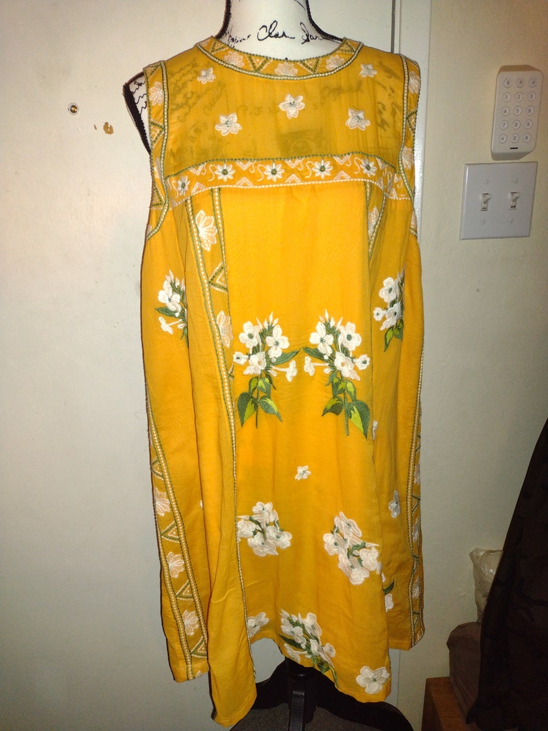 May include: A sleeveless yellow dress with white floral embroidery and green trim. The dress has a round neckline and a loose fit.