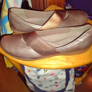 May include: A pair of brown leather shoes with a gold strap across the top. The shoes are sitting on a wooden surface.