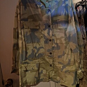 K&ouml;nnte beinhalten: Jacke mit Camouflage-Muster mit mehreren Taschen und Knopfverschl&uuml;ssen. Die Jacke hat einen Kragen und den Text "FREE PEOPLE" auf einem Etikett. Die Farbpalette umfasst Gr&uuml;n-, Blau- und Beiget&ouml;ne.