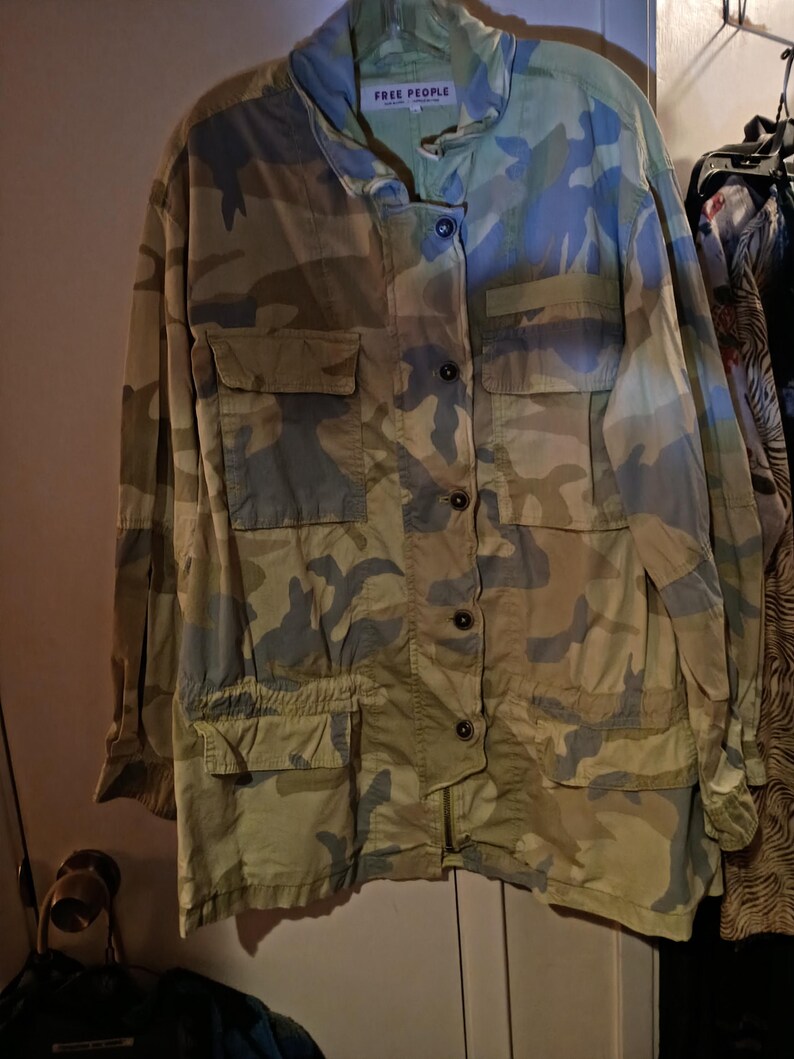 K&ouml;nnte beinhalten: Jacke mit Camouflage-Muster mit mehreren Taschen und Knopfverschl&uuml;ssen. Die Jacke hat einen Kragen und den Text "FREE PEOPLE" auf einem Etikett. Die Farbpalette umfasst Gr&uuml;n-, Blau- und Beiget&ouml;ne.