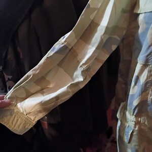 K&ouml;nnte beinhalten: Nahaufnahme eines &Auml;rmels einer Jacke mit Tarnmuster. Der &Auml;rmel ist beige, gr&uuml;n und blau und hat eine gekn&ouml;pfte Manschette. Die Jacke scheint aus einem leichten Stoff zu sein, der f&uuml;r w&auml;rmeres Wetter geeignet ist.