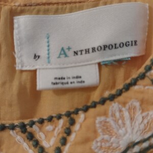 May include: A white tag with the text "by A+ Anthropologie" and "made in India/fabriqu&eacute; en Inde" on a yellow embroidered top.