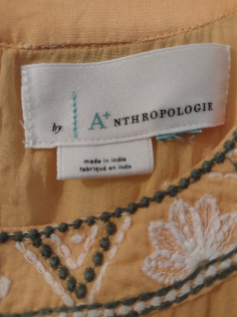 May include: A white tag with the text "by A+ Anthropologie" and "made in India/fabriqu&eacute; en Inde" on a yellow embroidered top.